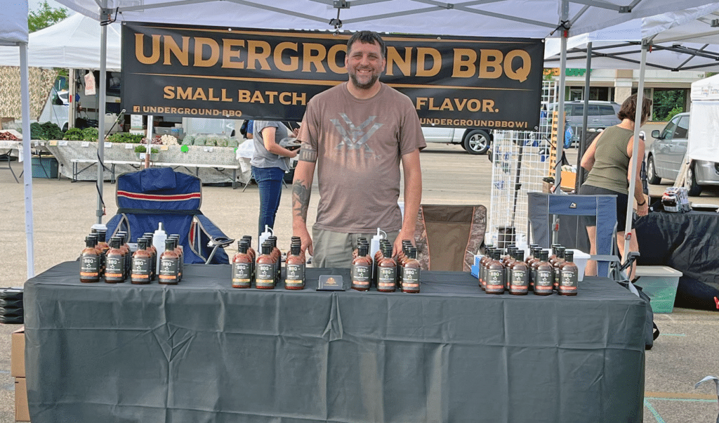 Man standing behind a table of BBQ sauce bottles at an outdoor market stall under an Underground BBQ banner.